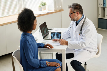 Obraz premium Mature doctor making notes in medical card and discussing the way of treatment with patient during her visit