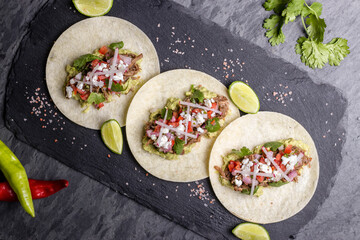 Mexican tacos carnitas with pulled meat, avocado, cilantro and chopped onions over a stone table.