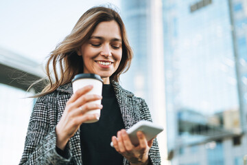 Young smiling woman in coat with coffee cup using mobile phone in evening city street