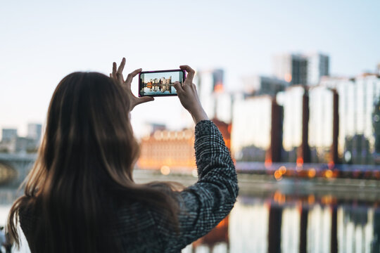 Young woman in coat taking photo on smartphone of evening urban city skyline