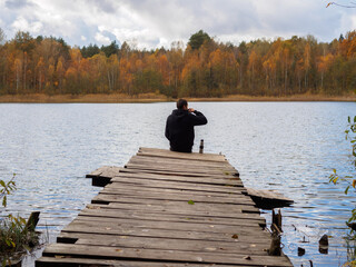 A young man is sitting on a wooden bridge by the lake and drinking tea, admiring the beauty of the autumn forest