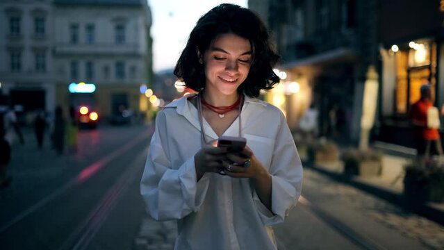 Smiling woman using smartphone on street with night city lights on background. Hipster female blogger typing text message on her cellphone
