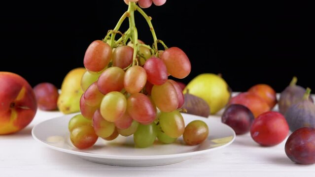 Close-Up, Female Hand Puts a Ripe Fresh Green Grape Bunch on a White Plate. Juicy berries of red, green color with drops of water on blurred background fruits on table. Healthy eating. Fall harvest.