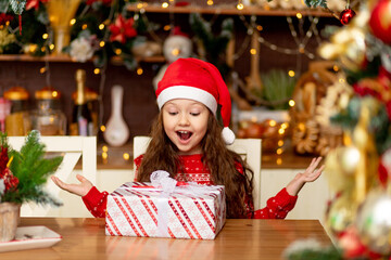 Fototapeta na wymiar cute little girl child in winter clothes Santa Claus hat and red sweater rejoices at the gift and laughs with happiness in the dark kitchen of the house, the concept of new year and Christmas