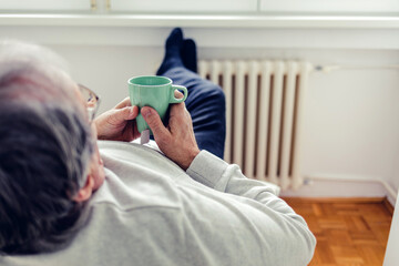 Man in warm sweater with hot tea sitting near radiator in winter. Energy crisis concept. Man with a...