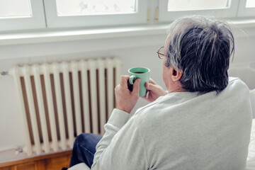 Man lying on a bed with woolen socks feet on the radiator heater. The man drinking hot tea,...