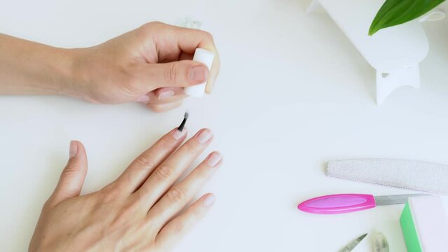 Manicurist woman applies nail gel polish on fingers. Female hands applying process gel manicure near ultraviolet lamp, bottles of nail polishes and tools - top view on white wooden table. Flat layout
