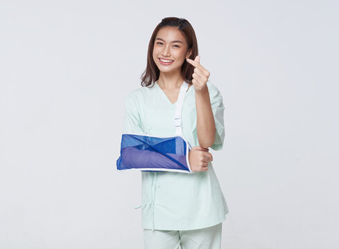 Asian teenage woman wearing a patient gown put on a cast due to injury from an accident showing hand mini heart with an impressive smile.insurance concept.