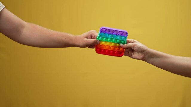 Male and female hands pulling colorful popular fidget toy in slow motion at yellow background. Unrecognizable Caucasian man taking away toy from woman at yellow background