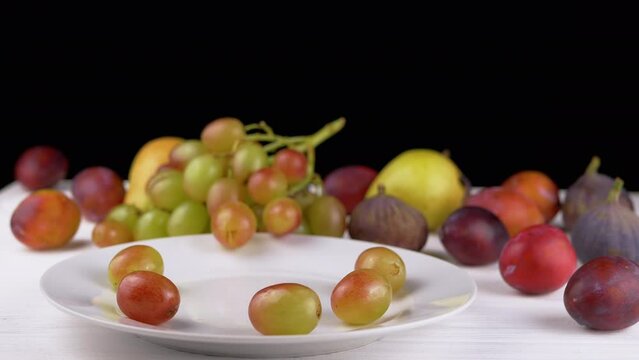 Female Hand Taking a Ripe Apple or Peach on a Plate with Grape and Fruit. Many different colorful fruits, plums, pears lie on the table. Harvest. Black and white background. Variety exotic products.
