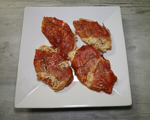 Four pieces of prosciutto-wrapped baked chicken on a white plater.