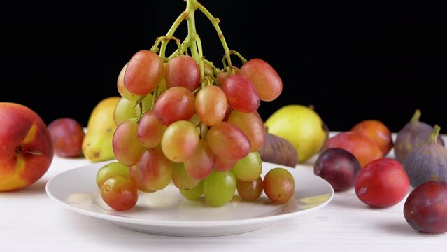 Close-Up, Female Hand Puts a Ripe Fresh Green Grape Bunch on a White Plate. Juicy berries of red, green color with drops of water on blurred background fruits on table. Healthy eating. Fall harvest.