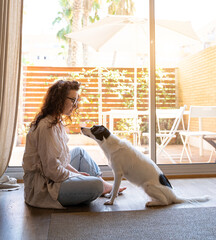 curly woman in glasses teaches dog commands. sitting on the floor opposite the window in the living room. Sunny daylight. dog attentively looking at face of owner. Pets devotion and attention
