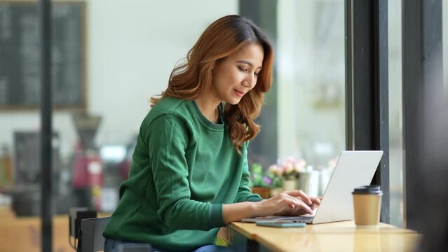 Cute young Asian woman sitting at cafe with her laptop happily. Asia female working on laptop computer at home.