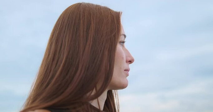 Red-haired young woman looks thoughtfully into the distance against a cloudy sky. Concept of appeasement, tranquility, peace, anxiety.