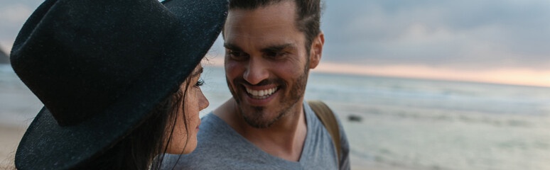 cheerful bearded man looking at woman in black hat on beach, banner.