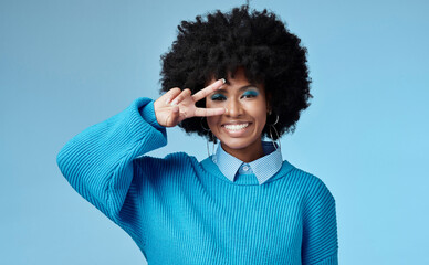 Young, black woman and shows peace sign with blue studio background, trendy style and afro. Smile,...