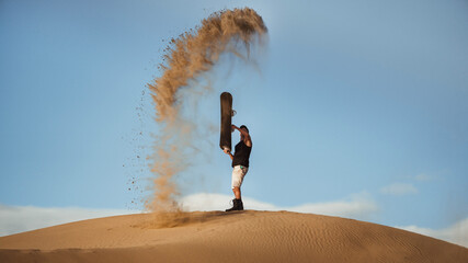 A man with a snowboard on a sand dune in the desert. The guy threw sand into the sky
