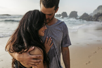 bearded man in grey t-shirt hugging happy girlfriend near ocean in portugal.