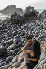 tattooed woman hugging happy bearded man on rocky beach in portugal.