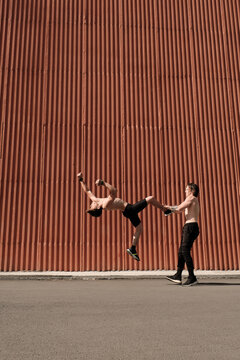 Vertical long shot of modern sporty man helping his friend to preform backflip during workout outdoors
