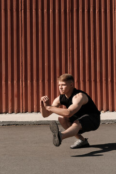 Vertical portrait of modern Caucasian man wearing black outfit doing one leg squat exercise outdoors