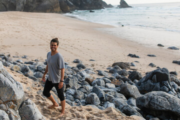 happy and bearded man in t-shirt standing near stones on beach near ocean.