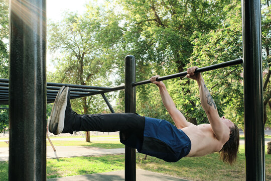 Horizontal shot of shirtless young Caucasian man with long hair exercising in local park on pull-up and dip bar on summer day