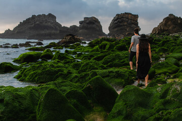 back view of tattooed woman in hat and dress standing behind man on mossy stones near ocean.