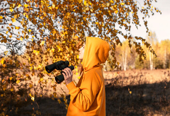 Young woman in yellow sweatshirt and hood with binoculars watching birds in the autumn forest Birdwatching, zoology, ecology Research, observation of animals Ornithology