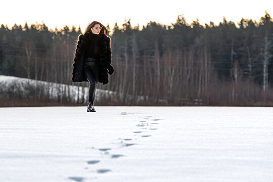 A Cheerful Girl In A Warm Fur Coat Walks Across The Open Field Leaving Footpath In The Snow