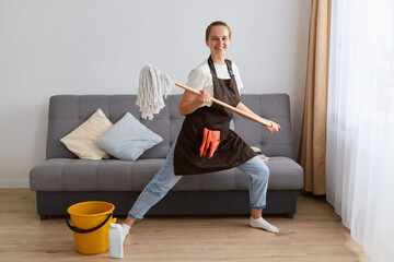 Fototapeta na wymiar Portrait of funny optimistic female washing floor with mop at home, doing domestic chores and having fun, dancing with cleaning tool, expressing positive emotions.
