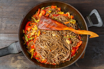 Soba with vegetables in soy sauce with spices in a frying pan with a wooden spatula on the table.