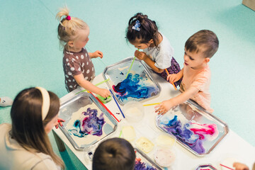 kids standing at the table full of watercolors and trying to paint with their teacher, nursery kids' concept. High quality photo