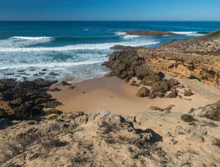 View of wild Rota Vicentina coast with ocean waves and red and orange sharp rock formations near Vila Nova de Milfontes, Portugal. Sunny day, blue sky