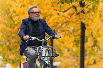 Cycling and active ways to spend time in autumn. Happy gray-haired bearded elegant man riding a city bike in beautiful park. Yellow fall trees in the background. High quality photo