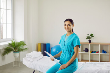 Portrait of smiling female masseuse or doctor in uniform hold patient card pose in cabinet in hospital. Happy young woman physiotherapist or therapist in clinic. Healthcare and medicine concept.