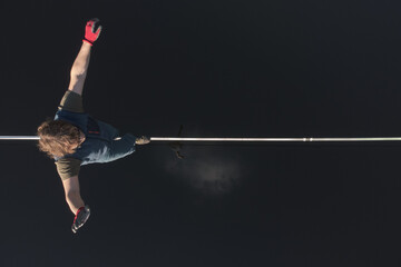 A male tightrope walker walks a slackline stretched over the mirror surface of the water. Drone...