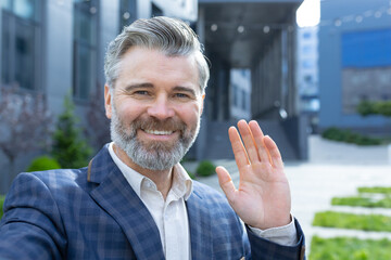 Video call of senior mature businessman, gray haired man looking phone camera and smiling, waving hand gesture of greeting, investor outside office building talking remotely with friends colleagues.