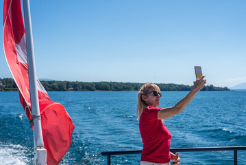 Mature blonde woman with glasses taking a selfie on a boat with Swiss flag fluttering on Lake Geneva.
