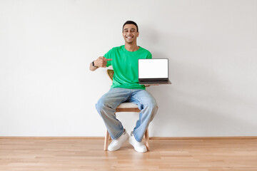 Cool website. Positive arab guy pointing at laptop with white empty screen, sitting on chair over white wall, mockup