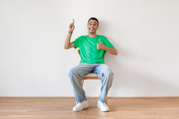 Cheerful arab man gesturing thumbs up and pointing finger up, sitting on chair over white wall and smiling to camera