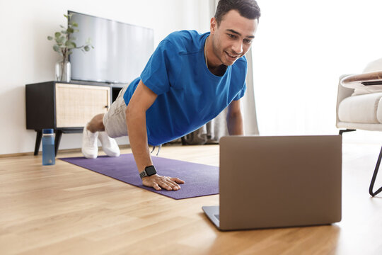 Young middle eastern man doing push-ups, having online fitness class on laptop from home, living room interior