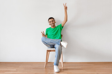 Emotional arab man sitting on chair over white wall, using modern wireless stereo headphones, enjoying music