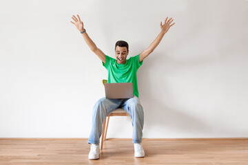 Overjoyed arab man in casual sitting on chair over white wall with laptop and raising hands up, got dream job