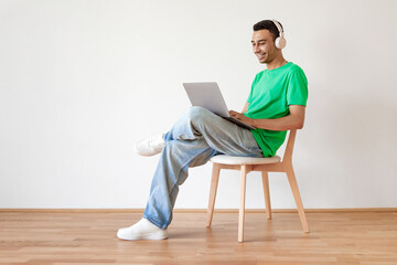 Young arab man using pc, working online on laptop and wearing wireless headphones, sitting on chair over white wall