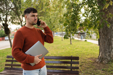 A young businessman outdoors in park sitting with his laptop talking on phone enjoying outside 