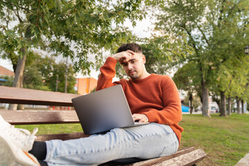 A young businessman outdoors in park sitting with his laptop talking on phone enjoying outside 