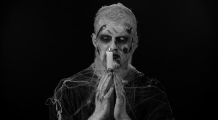 Frightening man with Halloween zombie bloody wounded makeup spells conjures over a candle. Horror...