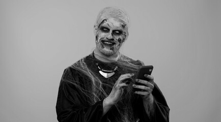 Frightening man with Halloween zombie bloody wounded makeup using mobile phone typing new post on web sms message, browsing, addiction of social networks. Sinister undead guy on gray wall background